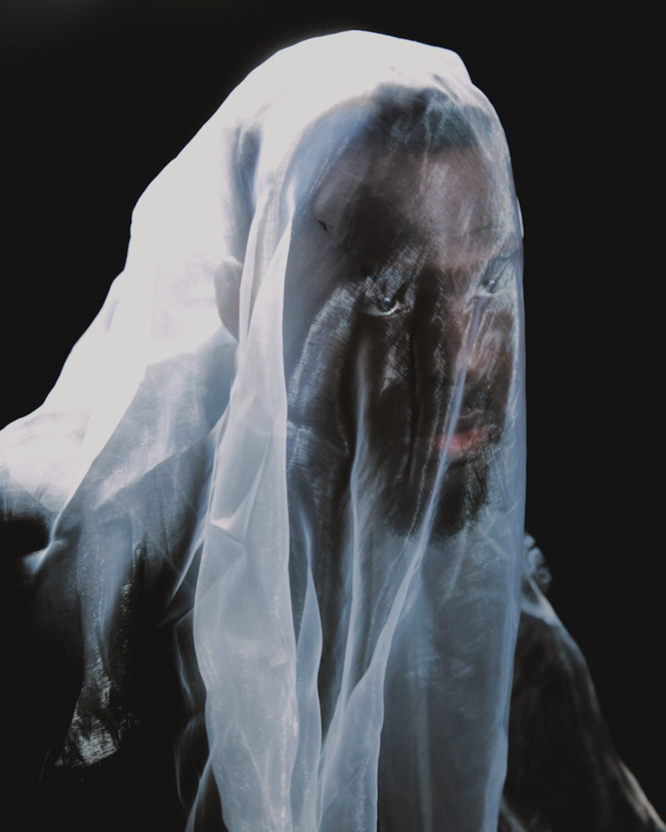 Varnish La Piscine looking at the camera through a translucent white veil draped over his head, against a black background.